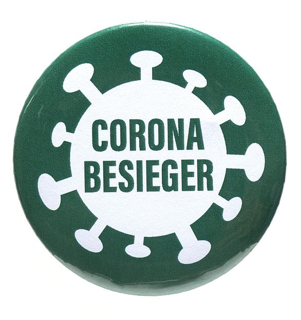 Ein runder Pin zum Anstecken, der den groben Umriss eines Virus andeutet, in dem der Text Corona Besieger in Großbuchstaben steht.