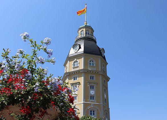 Der Schlossturm des Badischen Landesmuseums, auf dem die badische Flagge vor blauem Himmel weht. Im Vordergrund sind Blumen zu sehen.