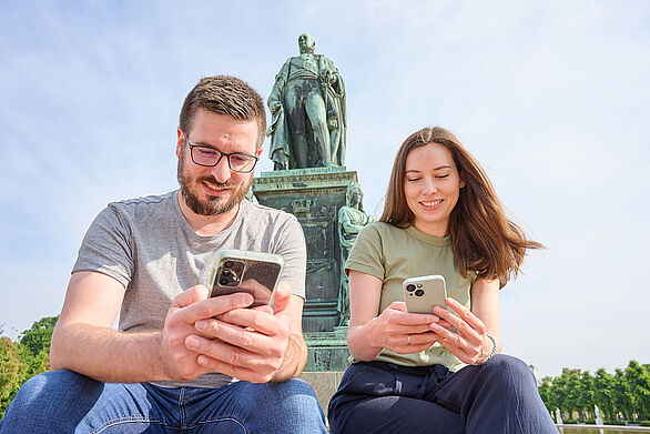 Zwei Menschen sitzen vor einem großen Denkmal und benutzen ihre Smartphones; der Himmel ist klar und sonnig.