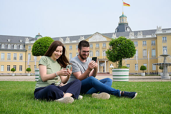 Zwei Personen sitzen auf einer Wiese vor dem Schloss Karlsruhe und nutzen ihre Smartphones. Im Hintergrund ist das historische Gebäude mit Fahne sichtbar.