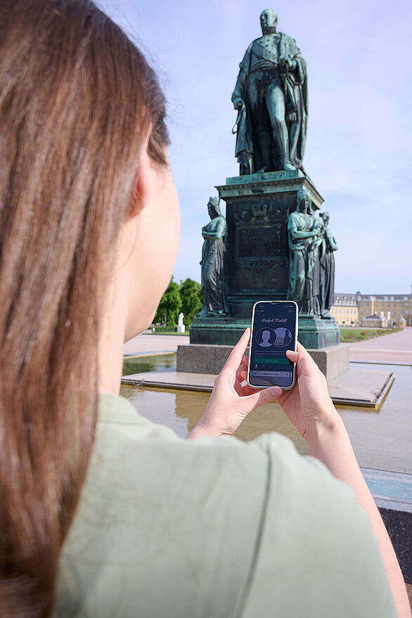 Frau mit braunen Haaren hält ein Smartphone mit geöffneter App und betrachtet ein historisches Denkmal bei Tageslicht.