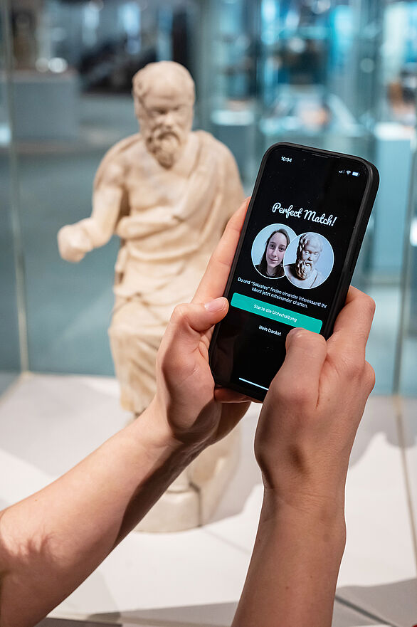 Zwei Hände nutzen die App Ping in einer Ausstellung. Im Hintergrund ist eine antike Skulptur eines bärtigen Mannes im Gewand.