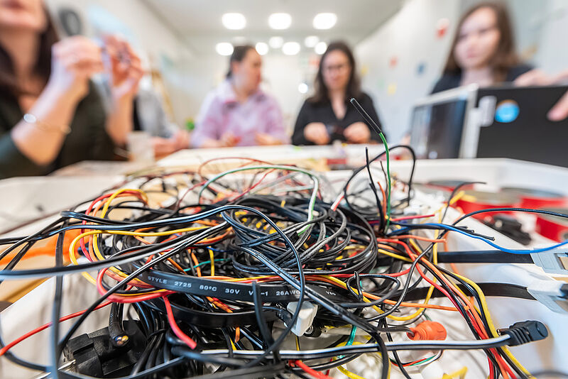 Bunte Elektrokabel liegen durcheinander auf einem Tisch im Hintergrund sind Menschen