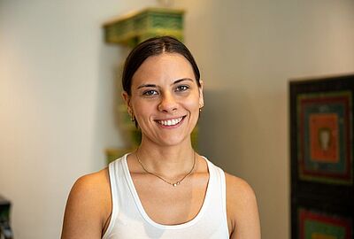 Porträt einer lächelnden Frau vor dem Hintergrund der Provenienzforschung im Museum beim Markt in Karlsruhe am Badischen Landesmuseum. Sie trägt ein weißes Top und eine feine Kette, während im Hintergrund ein farbenfrohes Kunstobjekt aus der Sammlung den Raum für Kultur und historische Aufarbeitung bereichert. Im Hintergrund ein Ofen der Art Deco Zeit.